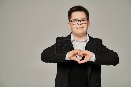 glücklicher Junge mit Down-Syndrom in Schuluniform und Brille mit Liebeszeichen mit Händen auf grau
