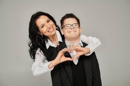 glückliche Frau in offizieller Kleidung zeigt Liebeszeichen in der Nähe ihres Sohnes mit Down-Syndrom in Schuluniform auf grau