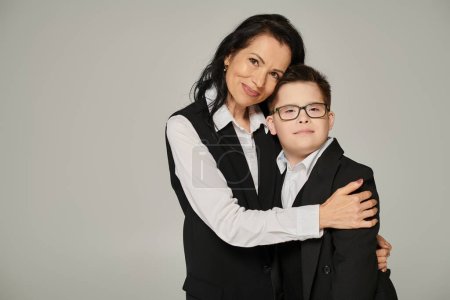 Téléchargez les photos : Joyeuse femme élégante embrassant son fils avec le syndrome du duvet en uniforme scolaire sur gris, amour inconditionnel - en image libre de droit