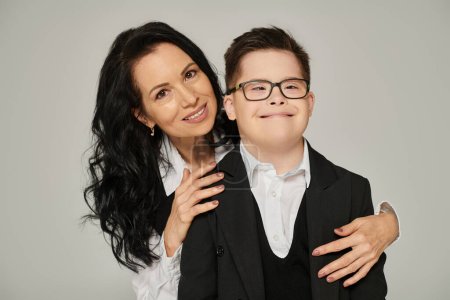 Téléchargez les photos : Femme d'âge moyen embrassant son fils avec le syndrome du duvet en uniforme scolaire tout en souriant à la caméra sur gris - en image libre de droit