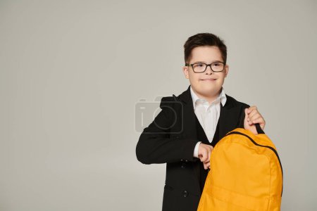 schoolboy with down syndrome holding yellow backpack and smiling on grey, inclusive schooling