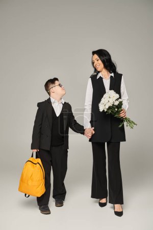 Téléchargez les photos : Écolier avec syndrome duvet tenant sac à dos jaune près de la mère avec des fleurs sur gris, pleine longueur - en image libre de droit