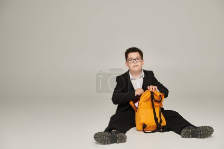kid with down syndrome in school uniform sitting with yellow backpack on grey and looking at camera