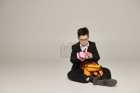schoolboy with down syndrome holding gift box and sitting near yellow backpack on grey, full length