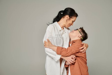 Téléchargez les photos : Garçon avec le syndrome du duvet et femme d'âge moyen en vêtements à la mode embrassant gris, famille unique - en image libre de droit