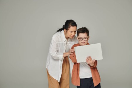 middle aged woman explaining online lesson to smiling son with down syndrome near laptop on grey