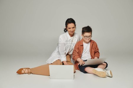 positive middle aged woman and boy with down syndrome sitting with laptops on grey, special family