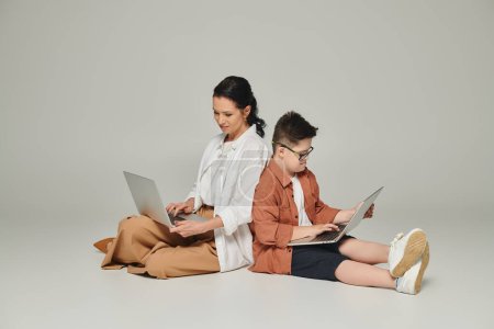 middle aged woman and kid with down syndrome sitting back to back with laptops on grey