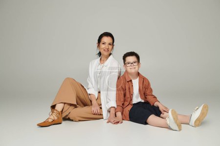 Téléchargez les photos : Joyeux garçon avec le syndrome du duvet et mère d'âge moyen dans des vêtements élégants souriant et assis sur gris - en image libre de droit