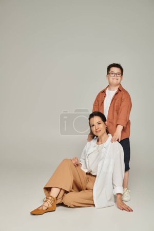 Photo for Happy boy with down syndrome in eyeglasses looking at camera near smiling mother sitting on grey - Royalty Free Image