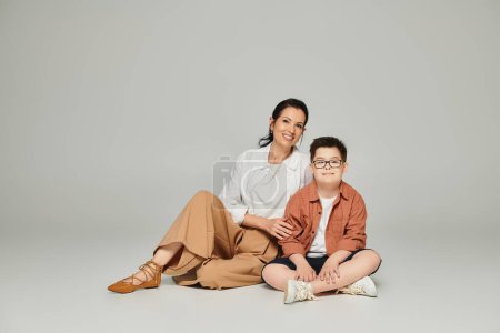 Téléchargez les photos : Femme d'âge moyen et garçon avec le syndrome du duvet assis et regardant la caméra sur gris, famille spéciale - en image libre de droit
