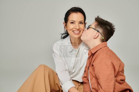 Photo for Boy with down syndrome kissing pleased mother looking at camera on grey, unconditional love - Royalty Free Image