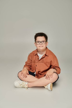 glücklicher Junge mit Down-Syndrom in trendiger Freizeitkleidung und Brille sitzend und lächelnd auf grau