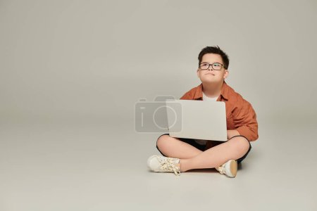 stilvoller und nachdenklicher Junge mit Down-Syndrom sitzt mit Laptop und gekreuzten Beinen auf grau