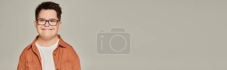 Photo for Cheerful kid with down syndrome, in eyeglasses and shirt looking at camera on grey, portrait, banner - Royalty Free Image