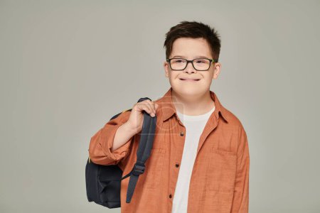 carefree schoolboy with mental disability holding backpack on grey, down syndrome concept