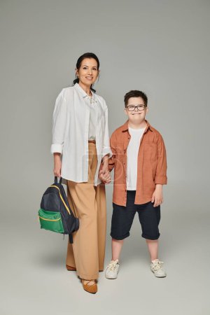 Téléchargez les photos : Femme d'âge moyen avec sac à dos scolaire et fils avec syndrome du duvet tenant la main sur gris, pleine longueur - en image libre de droit