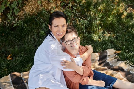 glückliche Frau mittleren Alters sitzt auf Decke im Park und umarmt Sohn mit Down-Syndrom, Qualitätszeit