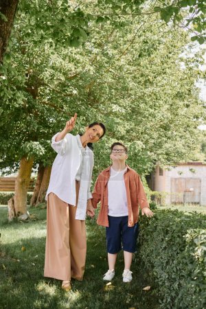 Téléchargez les photos : Femme d'âge moyen pointant du doigt pendant la sortie dans le parc avec un fils joyeux avec le syndrome du duvet - en image libre de droit
