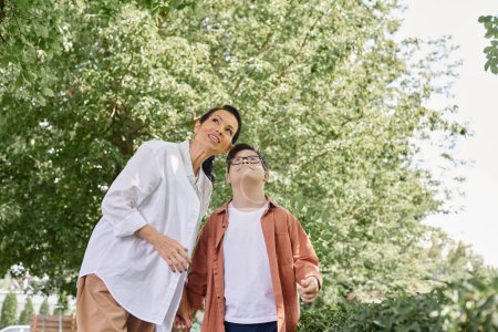 lächelnde Frau mittleren Alters und Junge mit Down-Syndrom, die im Park wegschauen, glückliche Momente