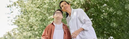 preteen boy with down syndrome near smiling mother looking away while walking in park, banner