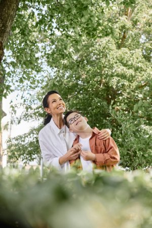 Foto de Alegre mujer de mediana edad y el hijo con síndrome de Down mirando hacia otro lado en primer plano borrosa en el parque - Imagen libre de derechos