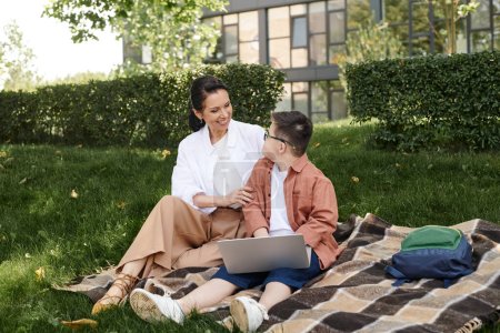 smiling woman and boy with down syndrome sitting near laptop on blanket in park, unique family