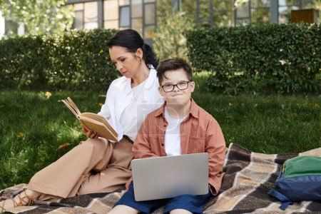 Téléchargez les photos : Femme d'âge moyen lecture livre près de fils avec le syndrome du duvet et ordinateur portable sur couverture dans le parc, loisirs - en image libre de droit