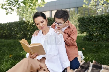 smiling middle aged woman reading book to son with down syndrome in park, quality time, leisure