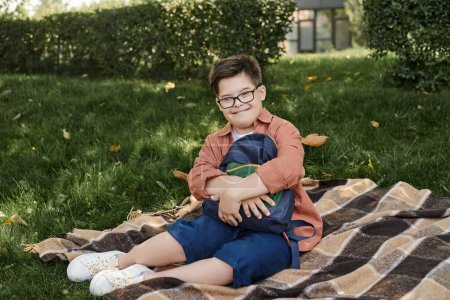 glücklicher und stilvoller Junge mit Down-Syndrom sitzt in der Nähe von Schulrucksack auf Decke im Park