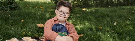 cheerful schoolboy with down syndrome, in eyeglasses, holding backpack on blanket in park, banner
