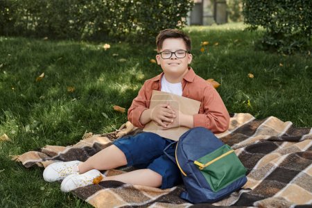 happy boy with down syndrome holding book while sitting near school backpack on blanket in park