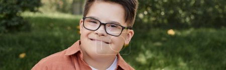 cheerful and genuine kid with down syndrome in eyeglasses smiling in park, portrait, banner