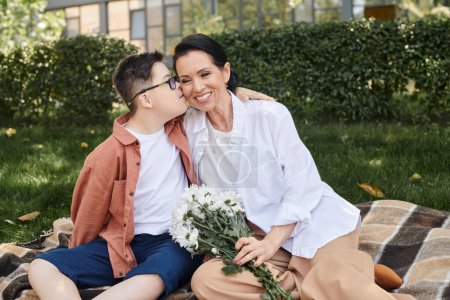 Téléchargez les photos : Enfant avec le syndrome du duvet embrasser mère assis avec des fleurs sur la couverture dans le parc, amour inconditionnel - en image libre de droit