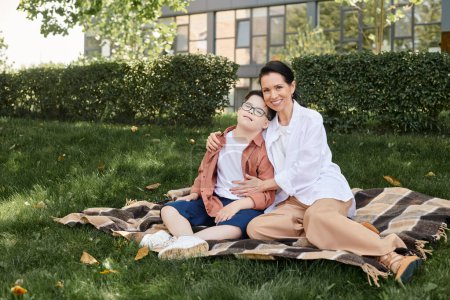 lächelnde Frau mittleren Alters, die ihren Sohn mit Down-Syndrom auf einer Decke im Park umarmt, Qualitätszeit