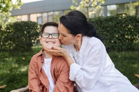 Frau mittleren Alters küsst unbeschwerten Sohn mit Down-Syndrom lächelnd mit geschlossenen Augen im Park