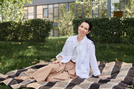 cheerful middle aged woman in trendy casual attire sitting on blanket in park and smiling at camera