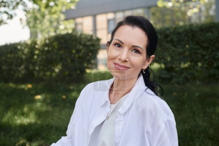 portrait of positive and stylish middle aged woman in white shirt smiling at camera in park