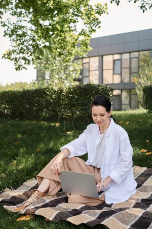 stylish and smiling middle aged woman working on laptop on blanket in park, work life balance