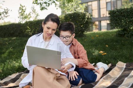 Foto de Feliz mujer de mediana edad sentado con el ordenador portátil cerca de hijo con síndrome de Down en la manta en el parque - Imagen libre de derechos