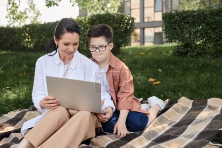 Téléchargez les photos : Enfant atteint du syndrome du duvet regardant un ordinateur portable près d'une mère souriante sur une couverture dans un parc, e-learning - en image libre de droit
