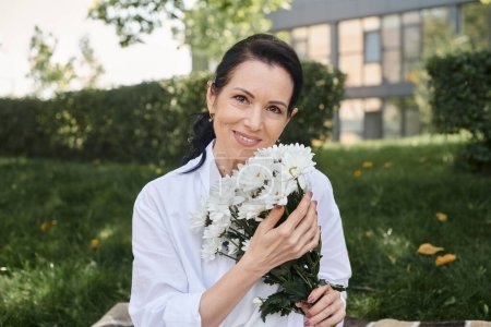 Téléchargez les photos : Portrait de joyeuse femme d'âge moyen tenant des fleurs et regardant la caméra dans le parc vert - en image libre de droit