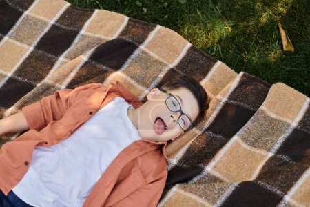 top view of carefree boy with down syndrome, in eyeglasses, sticking out tongue on blanket in park