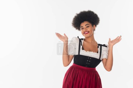 heureuse serveuse bavaroise afro-américaine en costume traditionnel invitant à l'oktoberfest sur blanc
