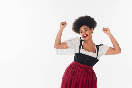 Téléchargez les photos : Excitée serveuse afro-américaine en costume bavarois traditionnel se réjouissant et gesticulant sur blanc - en image libre de droit