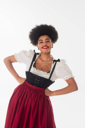 joyful african american oktoberfest waitress in traditional dirndl dress with hands on hips on white