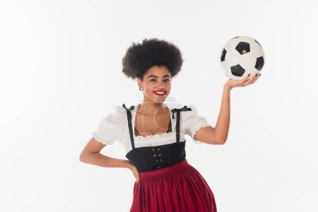 smiling african american bavarian waitress in dirndl with soccer ball on white, oktoberfest concept