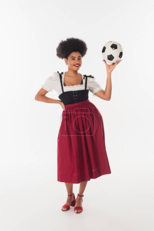 séduisante serveuse bavaroise afro-américaine en robe authentique posant avec ballon de football sur blanc