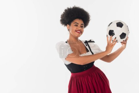 alegre octoberfest camarera en traje bavariano celebración de pelota de fútbol afroamericano en blanco