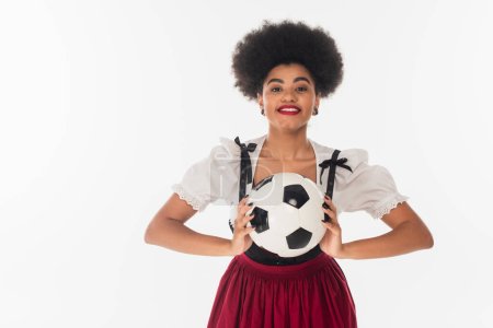 african american bavarian waitress in dirndl holding soccer ball on white, oktoberfest concept